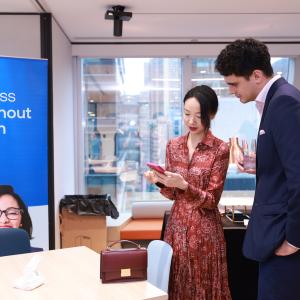 A woman in a patterned dress and a man in a suit discuss over a smartphone, with a VISTRA banner in the background.