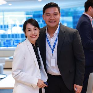 A man in a suit and a woman in a white blazer pose for a photo in a modern office with large windows and stylish furniture.