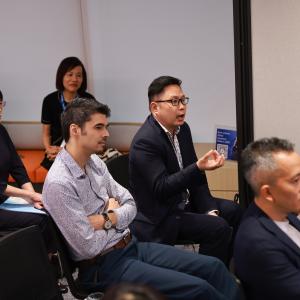A group of people attentively listen in a meeting room. One person gestures while speaking, conveying engagement. The atmosphere is professional and focused.