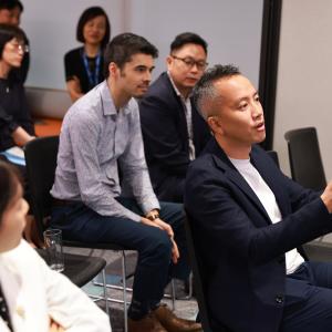 A group of people seated in a meeting room, attentively listening. A man in the foreground gestures expressively while speaking, conveying engagement.